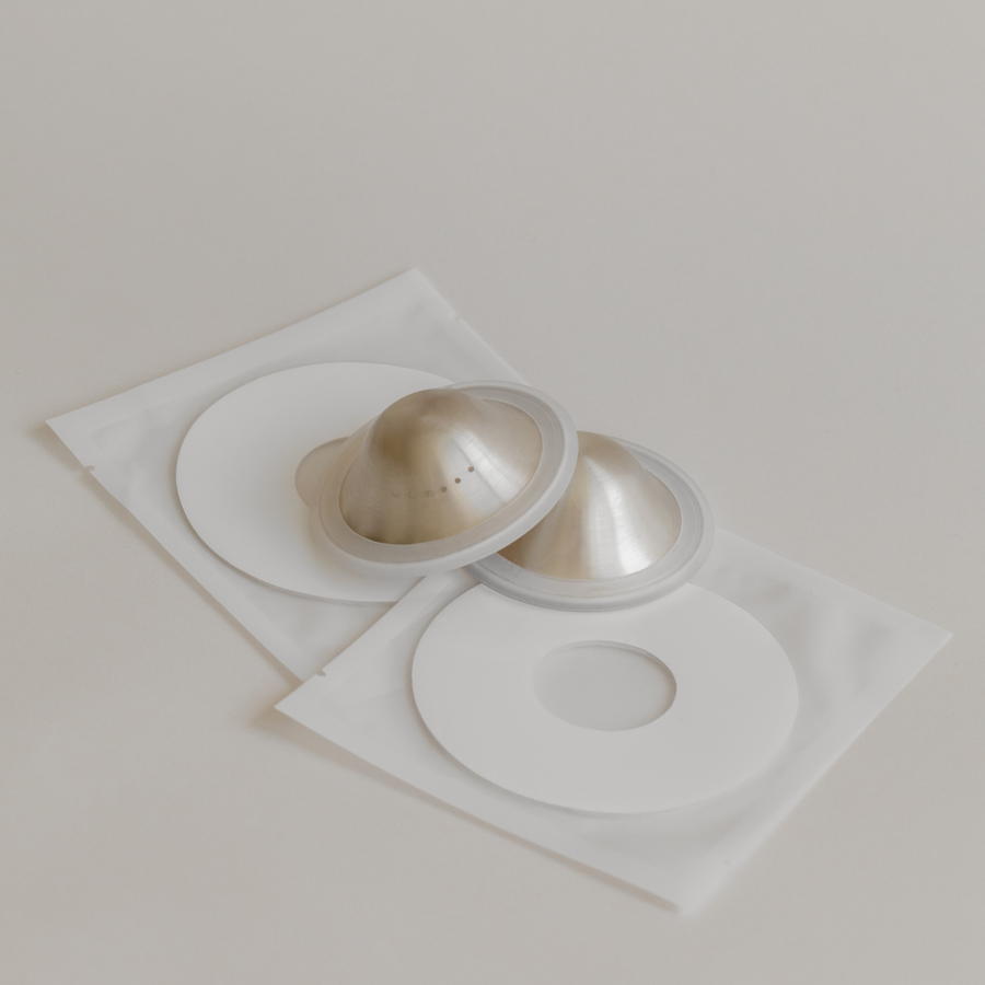 Two Bosom Ritual™ Discs beside their packaging on a beige surface, featuring breathability from Mammae The Embodied Mother.
