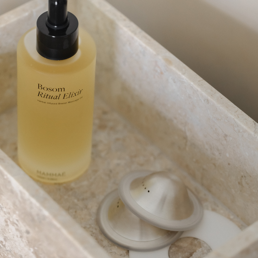 A bottle of Mammae The Embodied Mother's Bosom Ritual™ Objects on a marble counter next to a silver drain stopper.