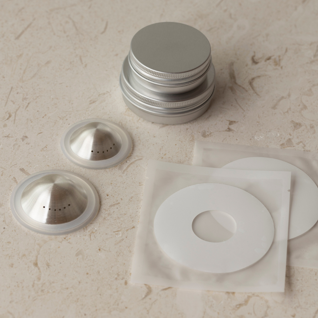 Mammae Silver Nipple Care Bundle placed on a light travertine table showing the silver nipple cups, hydrogel adhesives and aluminium hygiene storage vessel. 
