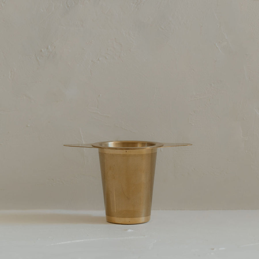 A small gold plated brass Ritualware Tea Infuser cup, equipped with a micro-mesh tea infuser, sitting on a table.