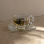 Mammae breastfeeding pyramid tea bag sitting in a vintage glass tea cup and saucer steeping in hot boiling water on a neutral background. 