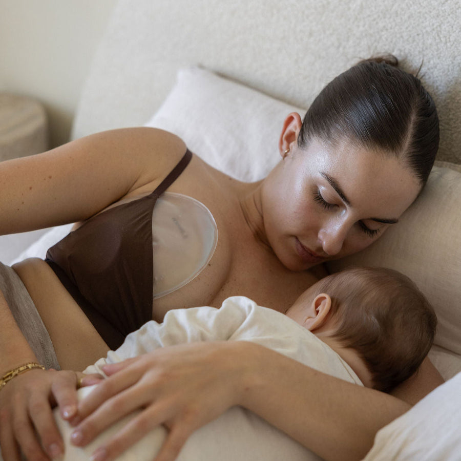 Mother breastfeeding a baby on a white linen bed in a cozy setting wearing the Mammae hot and cold breast care compress inside brown nursing bra. 