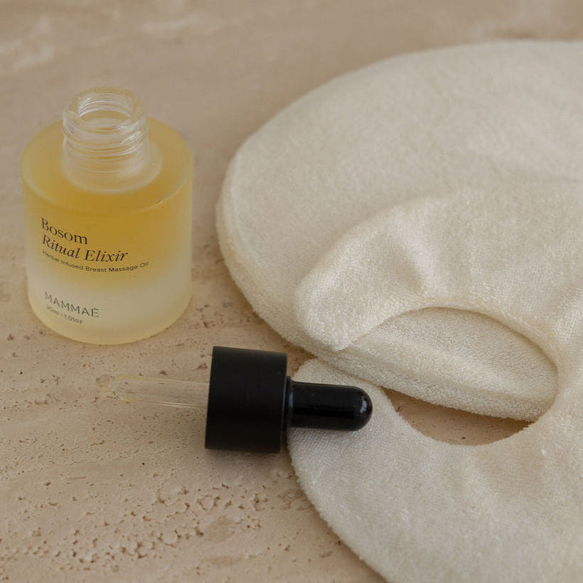 Mammae Bosom Ritual Elixir lymphatic breast  massage bottle with dropper on a beige surface. Mammae thermal therapy breast packs layered next to the bottle on a travertine surface. 