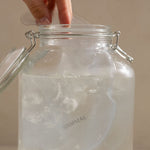 Clear glass jar with a lid on a neutral background and hand inserting the Mammae hot and cold breast care compress into iced water for lactation support. 