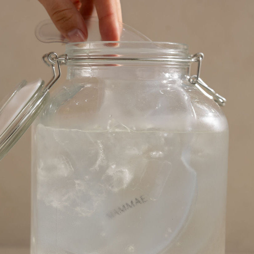 Clear glass jar with a lid on a neutral background and hand inserting the Mammae hot and cold breast care compress into iced water for lactation support. 