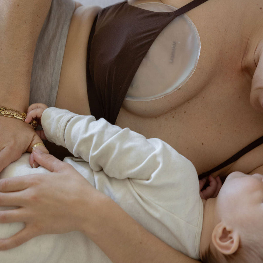 mother breastfeeding a baby in a home setting wearing the mammae hot and cold breast care compress inside of her brown nursing bra. 