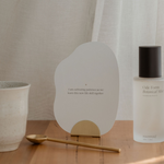 A bottle of Ode Form Botanical Mist Aromatherapy Ritual from the Mammae The Embodied Mother brand, promoting postpartum self-care and lactation support, on a table next to a cup of tea and the Ritualware Brass Teaspoon design to measure the Bosom Ritual Infusion Loose-lead Nursing Tea.