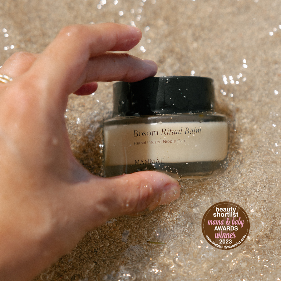 A hand is holding a glass jar of Bosom Ritual™ Balm in the ocean by Mammae The Embodied Mother, promoting breastfeeding, pumping and pregnancy wellness. Voted best nipple balm award winner at the mama and baby award 2023. 