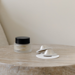 A jar of facial balm and two metallic Mammae The Embodied Mother Bosom Ritual™ Discs placed on a stone surface.
