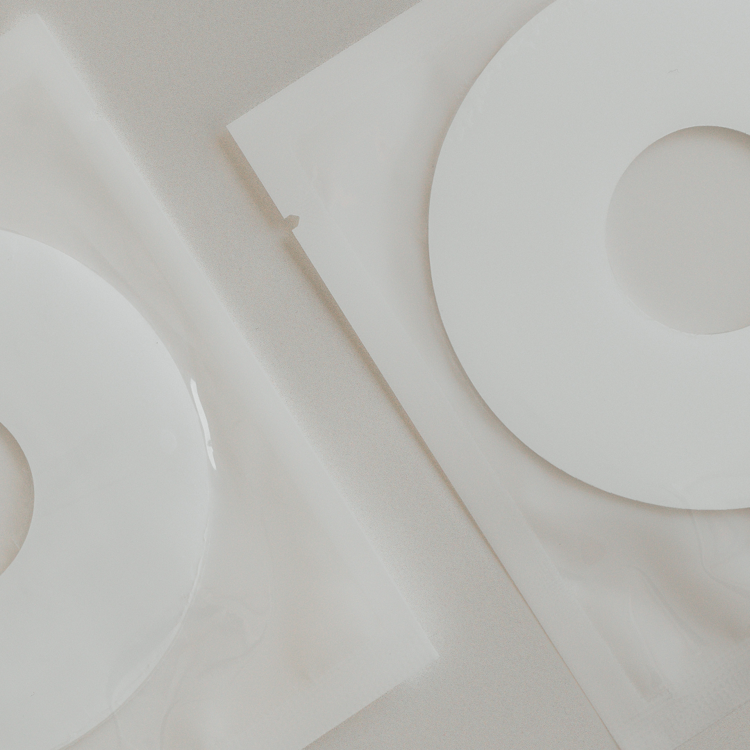 Overhead view of two white, sealed packaging bags with Aloe Infused Bosom Ritual Discs and circular cutouts against a gray background from Mammae The Embodied Mother.
