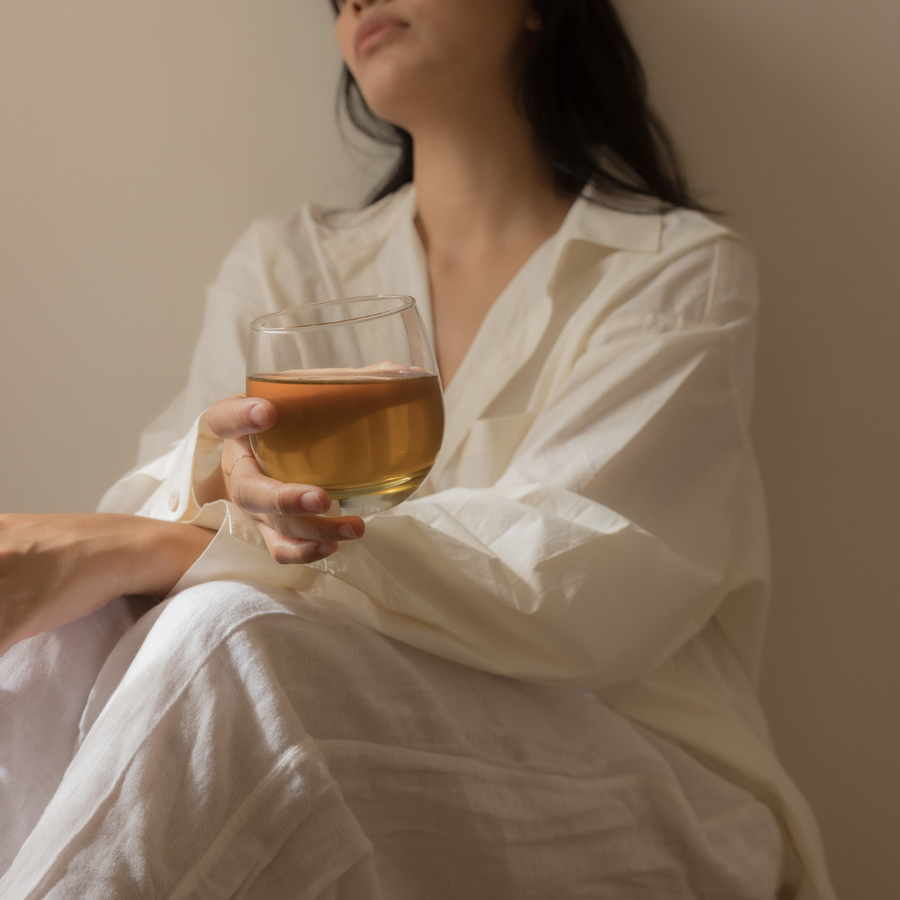 A breastfeeding mother dressed in white clothing holding a glass of Bosom Ritual™ Infusion certified organic nursing tea or breastfeeding tea formulated for postpartum mothers from Mammae The Embodied Mother.