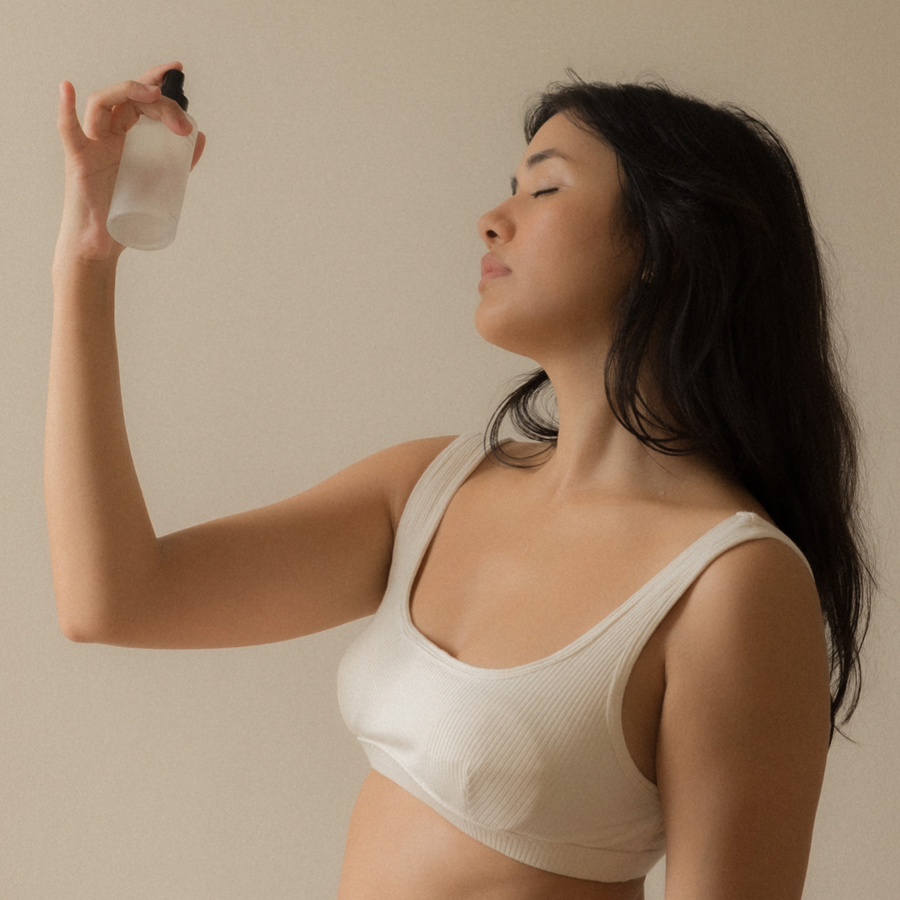 A woman in a white bikini holding the Mammae Botanical Mist spray bottle, promoting breastfeeding support.