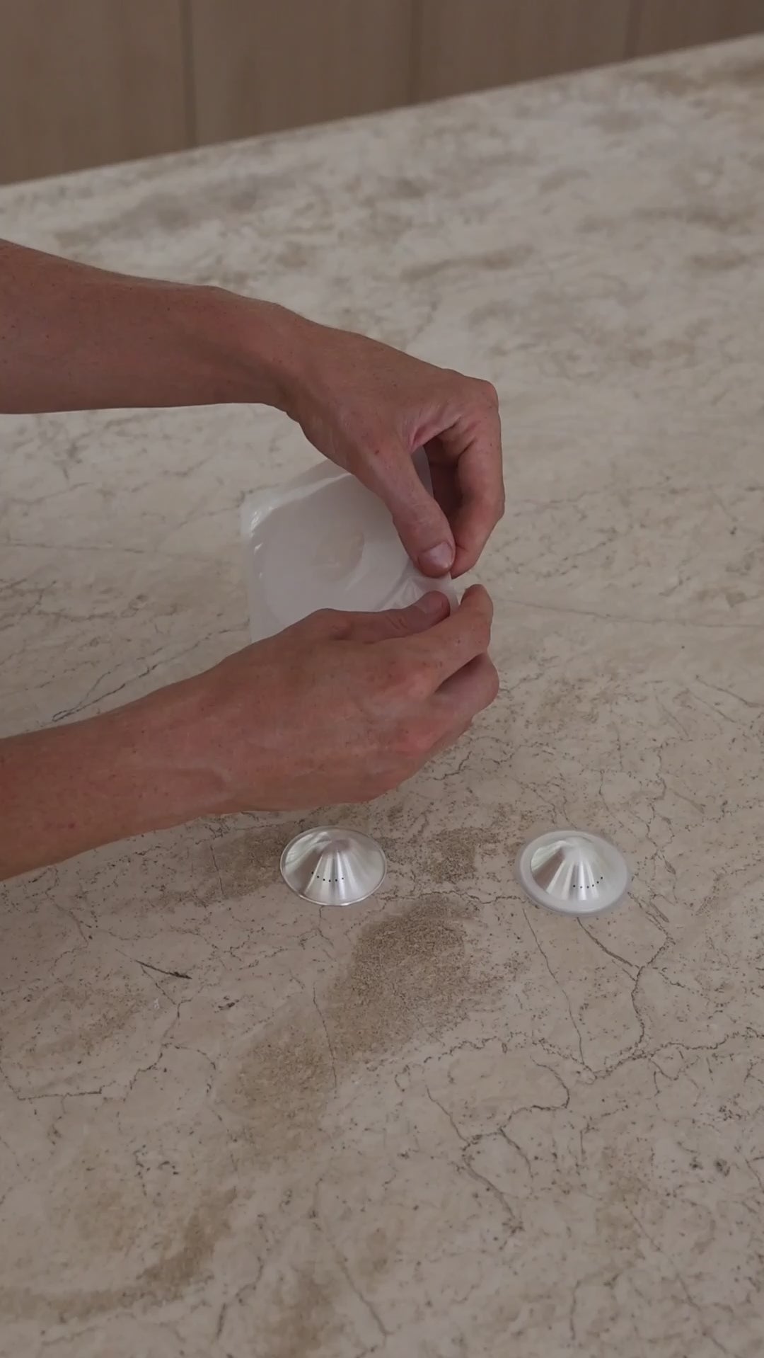 A new mother peeling back the Mammae aloe-infused hydrogel adhesives and sticking them down onto the surface of the breathable silver nipple cups on a travertine kitchen bench top. 