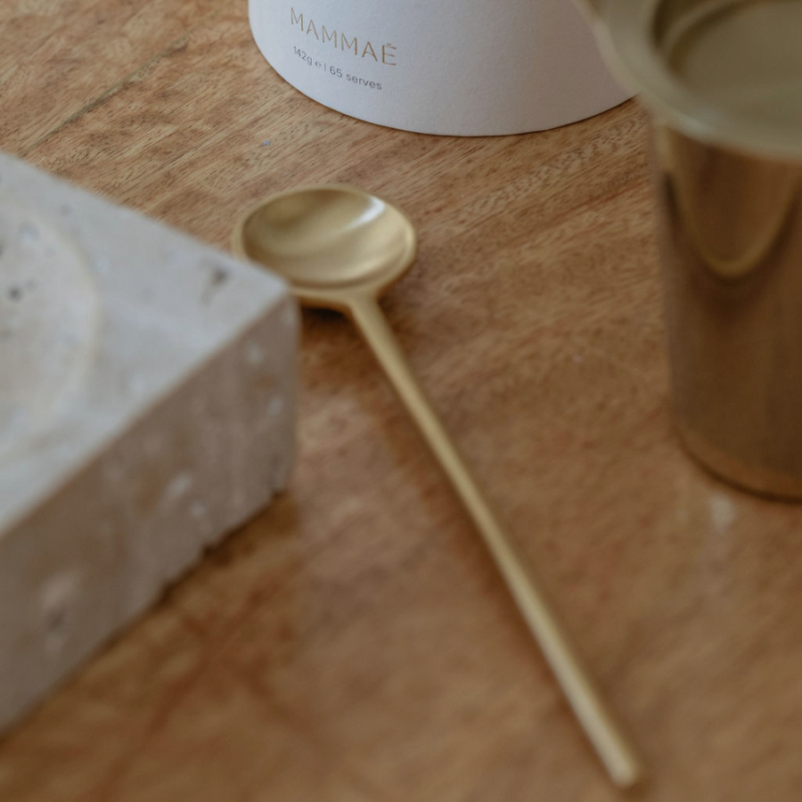 A Ritualware Brass Teaspoon from Mammae The Embodied Mother sits on a wooden table next to a cup, emphasizing its connection to postpartum and nursing stages.