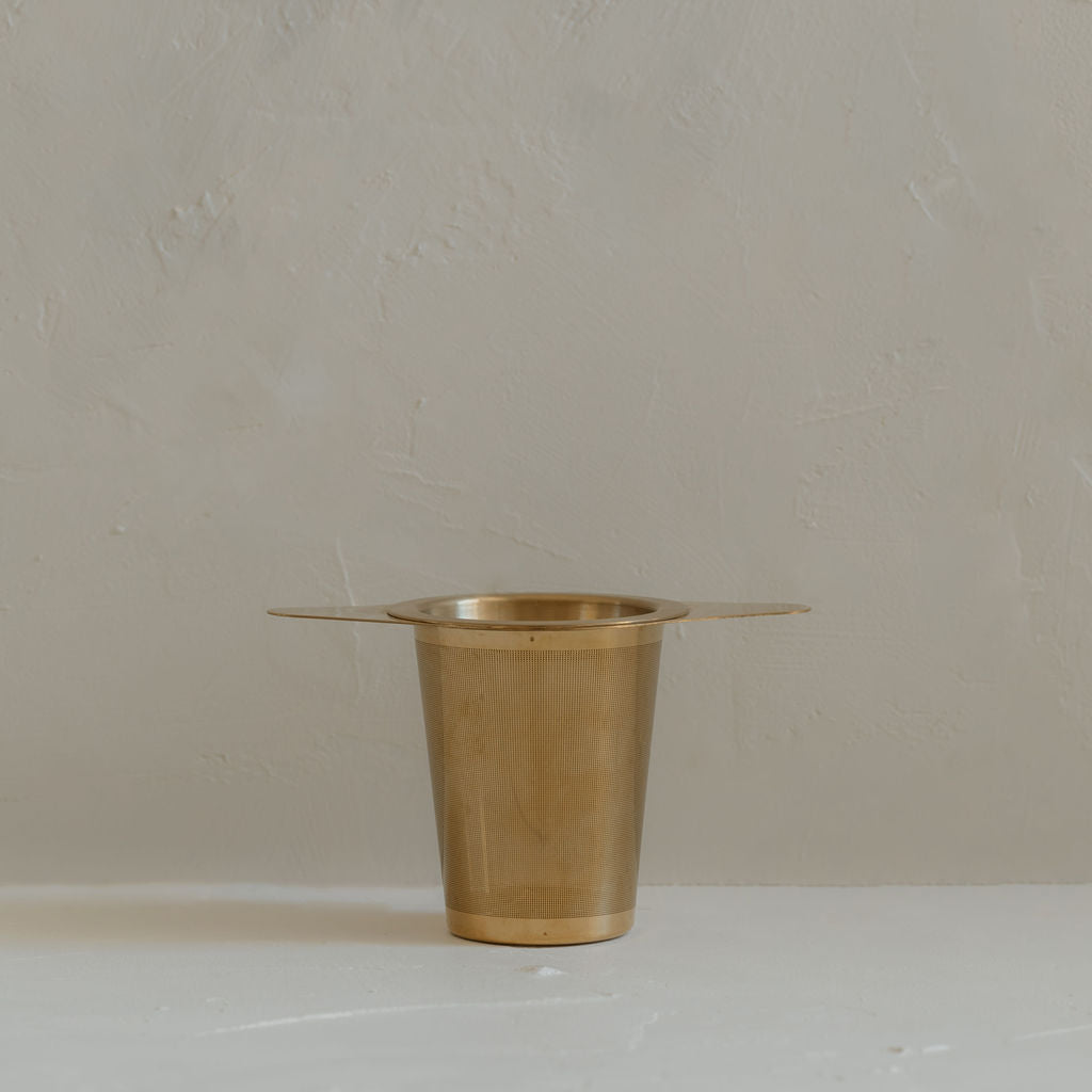 A small gold plated brass Ritualware Tea Infuser cup, equipped with a micro-mesh tea infuser, sitting on a table.