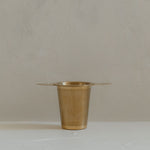 A small gold plated brass Ritualware Tea Infuser cup, equipped with a micro-mesh tea infuser, sitting on a table.
